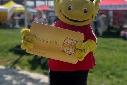 Das Ehrenamts-Maskottchen der Stadt hält eine große Ehrenamtskarte werbend in der Hand
