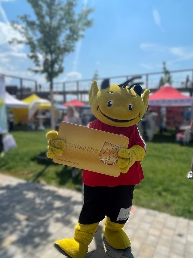 Das Ehrenamts-Maskottchen der Stadt hält eine große Ehrenamtskarte werbend in der Hand
