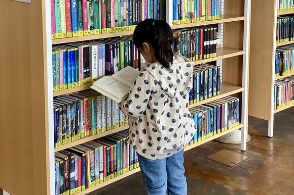 Ein Mädchen steht mit einem aufgeschlagenen Buch in den Händen vor einem Bücherregal.