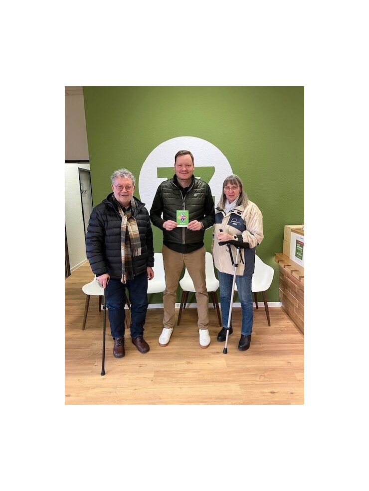 Drei Personen stehen in der Therapiezeit Opladen, der mittlere hält das Barrierefrei-Siegel in der Hand